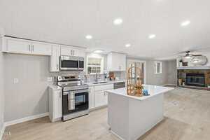 A modern kitchen with white cabinets, stainless steel appliances, an island with a basket tray, light wood flooring, and a view into a living area with a fireplace and ceiling fan.