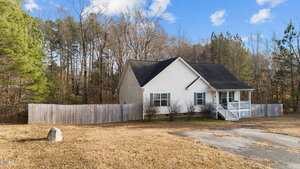 A white single-story house with a front porch, black roof, and steps, sits in front of a wooden fence. The yard is mostly brown grass, bordered by wooded trees under a blue sky with scattered clouds.