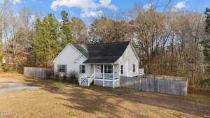 A single-story white house with a dark roof, front porch, and fenced backyard sits on a large, grassy lot. Leafless trees and pine trees surround the property under a partly cloudy sky.