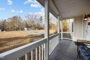 A view from a covered front porch with white railings, looking out over a dry lawn and driveway toward a distant house, with leafless trees and a partly cloudy sky in the background.