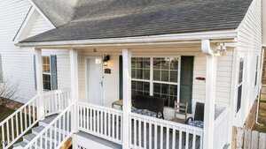 A white house with a covered front porch featuring a white railing, a light blue door, two green window shutters, outdoor seating, and steps leading down to the yard.