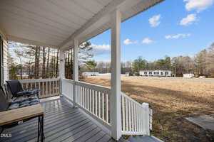 Covered front porch with white railings, two cushioned chairs, and a small table overlooks a large grassy yard with trees and a white building in the distance under a blue sky with scattered clouds.
