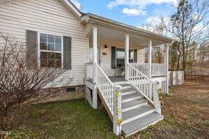 A white single-story house with a covered front porch, gray steps, white railings, black window shutters, and a small outdoor table with two chairs. The yard has patchy grass and leafless bushes.