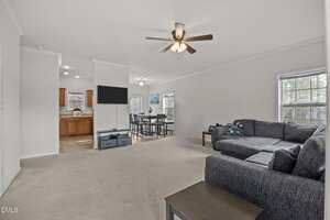 A spacious living room with beige carpet, a gray sectional sofa, a ceiling fan with lights, a flat-screen TV, and large windows. The space opens to a dining area and kitchen with wooden cabinets.