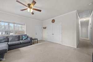A living room with beige carpet, a gray sectional sofa, a small side table, a ceiling fan with lights, a double door closet, a window, and a hallway leading to other rooms.