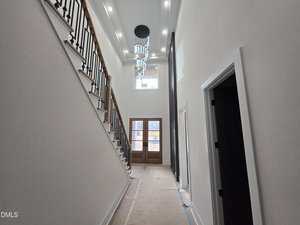 A modern, well-lit entry hallway with high ceilings, a large chandelier, wooden double front doors, and an open staircase with black railings on the left side. The floor is covered for protection.