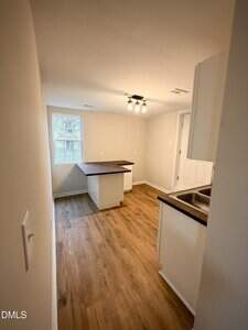A small kitchen with wood-style flooring, white walls, a window, brown countertops, and a ceiling light fixture. There is a counter area with an L-shaped layout and an under-mount sink on the right side.