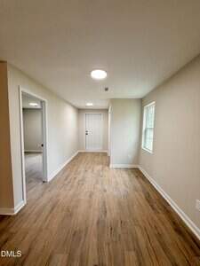 A narrow hallway with wood-look flooring, beige walls, white trim, two windows on the right, open doorways on the left, and a white entry door at the far end. Ceiling lights are installed overhead.