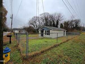 A small single-story house with white siding and a gray roof sits behind a chain-link fence on a grassy lot. A blue and yellow fire hydrant is near the fence, and utility lines run overhead on a cloudy day.