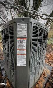 Close-up view of a large outdoor air conditioning unit with warning and information labels, surrounded by wet leaves and a chain overhead. Trees and overcast sky are visible in the background.