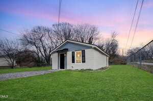 A small, single-story white building with a gray roof sits on a grassy lawn at sunset at 636 Flint Street in Henderson. The building features a black door and window shutters, with bare trees and a chain-link fence in the background.