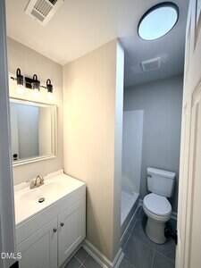 A small bathroom with a white vanity, sink, and mirror on the left, gray tile floor, a white toilet, and a shower area separated by a partial wall. Ceiling lights and neutral wall colors are present.