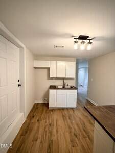 A small kitchen area with white cabinets, a stainless steel sink, brown countertops, wood-look flooring, and a ceiling light fixture. A white door is on the left, and a hallway is visible in the background.