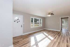 Empty living room with light gray walls, wood-style flooring, a white front door, two windows letting in sunlight, a ceiling fan with lights, and an open doorway leading to another room.