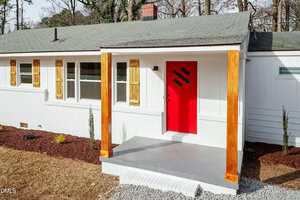 A single-story white house with a gray roof, light wood shutters, a bright red front door with three black slats, and a small covered porch with wood posts. The yard has brown mulch and small plants.