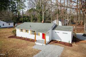 A single-story white house with a green roof at 802 Roanoke Avenue, Henderson features a red front door, attached garage, small front porch, and gravel driveway. The yard is surrounded by trees with neighboring houses in the background.