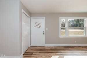 A white door with a round black handle is set in a beige wall next to a double closet. A window on the right shows a lawn and trees outside. The floor is light wood, and the room is empty.