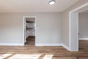 A room with light gray walls, white trim, and wood-style flooring. There is an open doorway leading to a white kitchen on the left and another open entry to a separate room on the right. A ceiling light is visible.