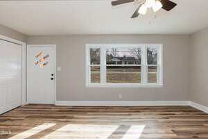 A bright, empty room with light gray walls, wood-look flooring, a ceiling fan with lights, a white door with three diagonal glass panels, double closet doors, and a large window facing the street.