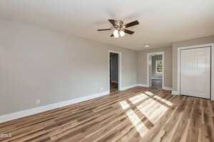 A spacious, empty room with light gray walls, wood-patterned floors, a ceiling fan with lights, two doorways, and a white closet door. Sunlight streams in through a window that is not visible in the image.