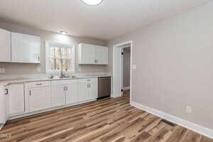 A modern kitchen with white cabinets, stainless steel appliances, a sink under a window, marble countertops, and light wood flooring. The walls are painted light gray and the space appears clean and empty.