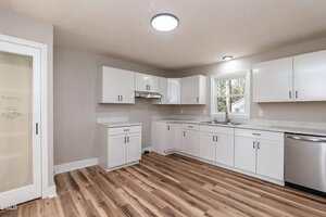 A modern kitchen with white cabinets, stainless steel appliances, marble countertops, wood flooring, a window above the sink, and a frosted glass pantry door on the left side.