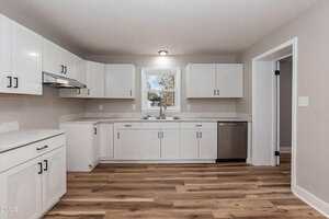 A modern kitchen with white cabinets, marble countertops, stainless steel dishwasher, single window above a double sink, and wood-style vinyl flooring. Walls are light beige and appliances are minimal.