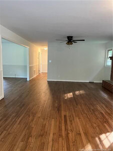 A spacious room with hardwood floors, white walls, a black ceiling fan, and a partial view of a brick fireplace. An open doorway leads to another room with matching flooring and wainscoting.