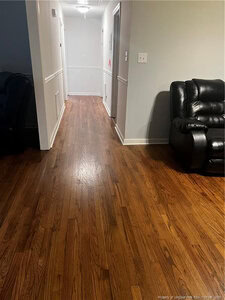 A hallway with wood flooring, white walls with trim, and a black leather recliner on the right. The hallway is well-lit and leads to a door at the end.