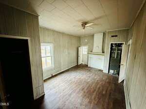 A small, empty room with wood-paneled walls, a wood floor, a ceiling fan, two doors, a window, and a wall-mounted cabinet next to a white fireplace. The room appears to be in an older house.