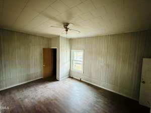 An empty room with wood-paneled walls, a window, an open door, white ceiling tiles, a ceiling fan, and a worn wooden floor. The room appears to be unfurnished and in need of renovation.