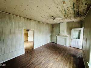 An empty room with wood-style flooring, paneled walls, a white fireplace, and a ceiling fan. The ceiling shows signs of peeling paint and water damage. There are two doorways, one with a glass door and one open.