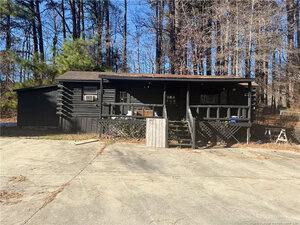 A small black log cabin with a covered front porch sits in front of a wooded area. The house has a slanted roof, steps leading to the porch, and is bordered by tall, leafless trees. A concrete driveway is in the foreground.