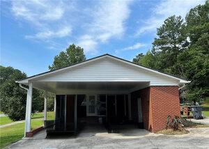 A single-story house with a red brick exterior and white siding features a covered carport. Trees and grass surround the home, and a driveway leads up to the carport. The sky is mostly clear with a few clouds.