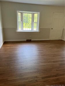 An empty room with hardwood floors, light-colored walls, a white door, and a large bay window letting in natural light. There is a floor vent beneath the window and minimal decorative features.