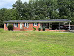 A single-story red brick house with black shutters, a white front door, and a covered carport attached on the right side sits at 1675 US 15 in Creedmoor, surrounded by a large grassy yard with trees in the background.