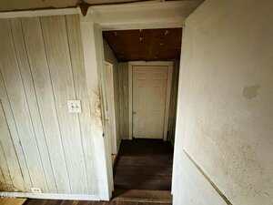 A narrow hallway with worn wood-paneled walls, dirty surfaces, and a closed white door at the end. The ceiling shows signs of damage, and the area appears dimly lit and in poor condition.