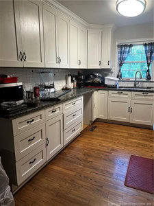 A kitchen with white cabinets, black handles, granite countertops, and a stainless steel sink under a window with a curtain. Various appliances and dishes are on the counters, and the floor is wooden with a red mat.