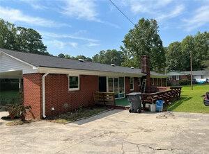 A single-story red brick house with a black shingle roof, an outdoor brick chimney, and a concrete driveway. There are trash bins and blue bags near a wooden railing. Trees and another house are in the background.
