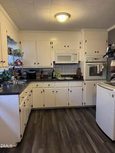 A kitchen with white cabinets, a microwave, an oven, black countertops, a dish rack, a coffee maker, and a standalone white freezer on a wood-patterned floor under a ceiling light.