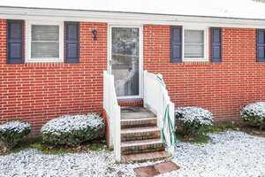 A front entrance to a red brick house with a white storm door, white railing, three steps, a green garden hose, two windows with blue shutters, and bushes dusted with snow.