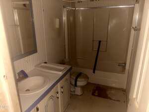 A bathroom with a bathtub-shower combo, toilet, and sink. The toilet and tub are taped shut, and there are stains on the floor. The room has wallpaper, a mirror, and worn cabinets. The lighting is dim.