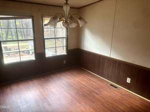 Empty room with wood-style laminate flooring, dark wood paneling on the lower walls, light wallpaper on the upper walls, a small chandelier-style light fixture, and two windows showing an outdoor view.