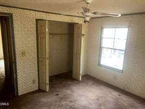 A small empty room with floral wallpaper, a ceiling fan, an open closet with double doors, a window letting in daylight, and worn carpet flooring. An open doorway leads to another room.