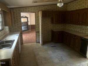 A dated kitchen with brown wood cabinets, beige countertops, floral wallpaper, a double sink under a window, dishwasher, ceiling fan, and an open doorway leading to a living area with wood flooring.