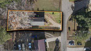 Aerial view of a rectangular property outlined in orange, containing a house, a driveway, and trees. The property is bordered by streets on two sides, with parked cars and other houses nearby.