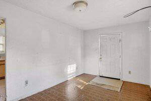 An empty room with white walls, a ceiling light, a door with a window, and a worn-out floor with visible stains and patches. Part of a kitchen with wooden cabinets is visible through an open doorway on the left.