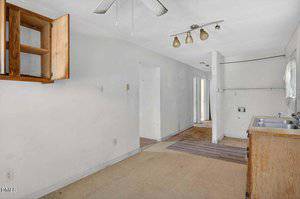 A worn, empty kitchen with light-colored walls, an open wooden cabinet, a double sink, basic counters, visible plumbing connections, and a ceiling fan. The room leads to a hallway with multiple doorways.