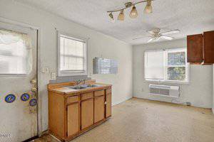 A small, worn kitchen with a double sink, wooden cabinets, and old linoleum flooring. There is a wall air conditioning unit under two windows and a ceiling fan with a light fixture above.
