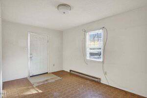 Empty room with white walls, a white ceiling light, a white door, a small window with blinds, exposed cords, a wall heater, and brown patterned flooring. The space appears unfinished or in need of renovation.
